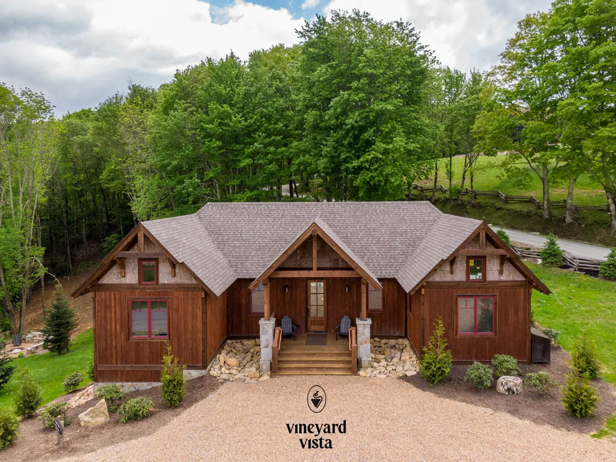 The image shows the charming wooden facade of the Vineyard Vista cabin, set among lush landscaping and mature trees. Stone steps lead up to a welcoming porch, ideal for sunset views and stargazing. Vibrant blooms and greenery surround the home, hinting at the beauty of each season in this tranquil mountain setting.