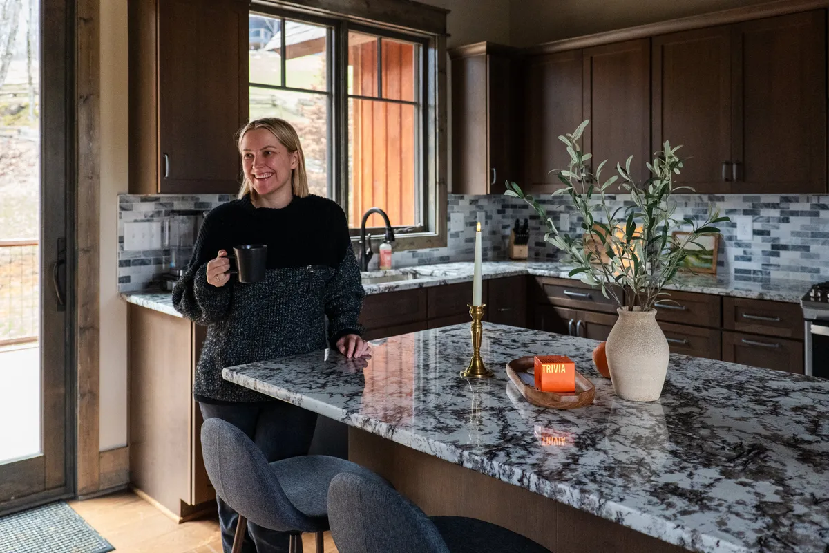 A close-up of the kitchen island, with a woman enjoying a mug of coffee, granite countertops, and a window pouring in daylight. The inviting kitchen ambiance is further enhanced by the visible appliances and relaxed mood.