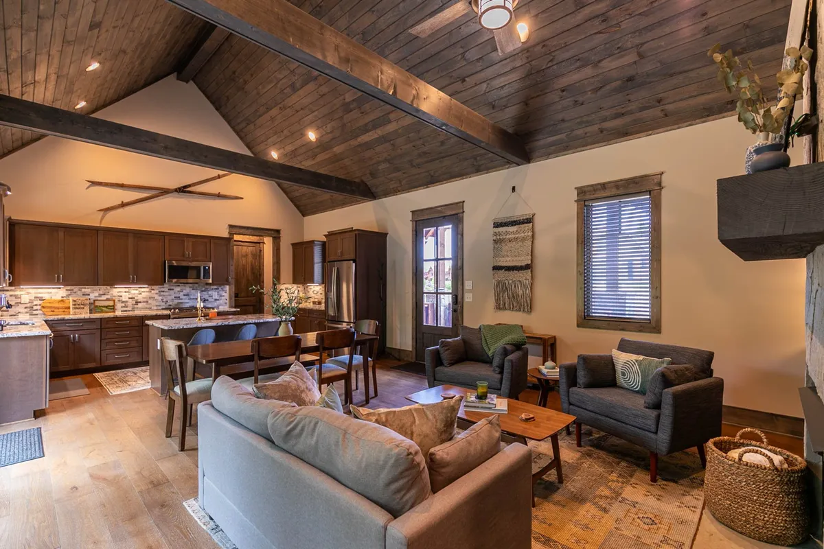 A wide view of an open-plan interior, including a fully stocked kitchen with visible appliances, a wooden dining table, exposed beams, and a relaxed seating area. The space is bathed in natural light, highlighting the communal and inviting atmosphere.