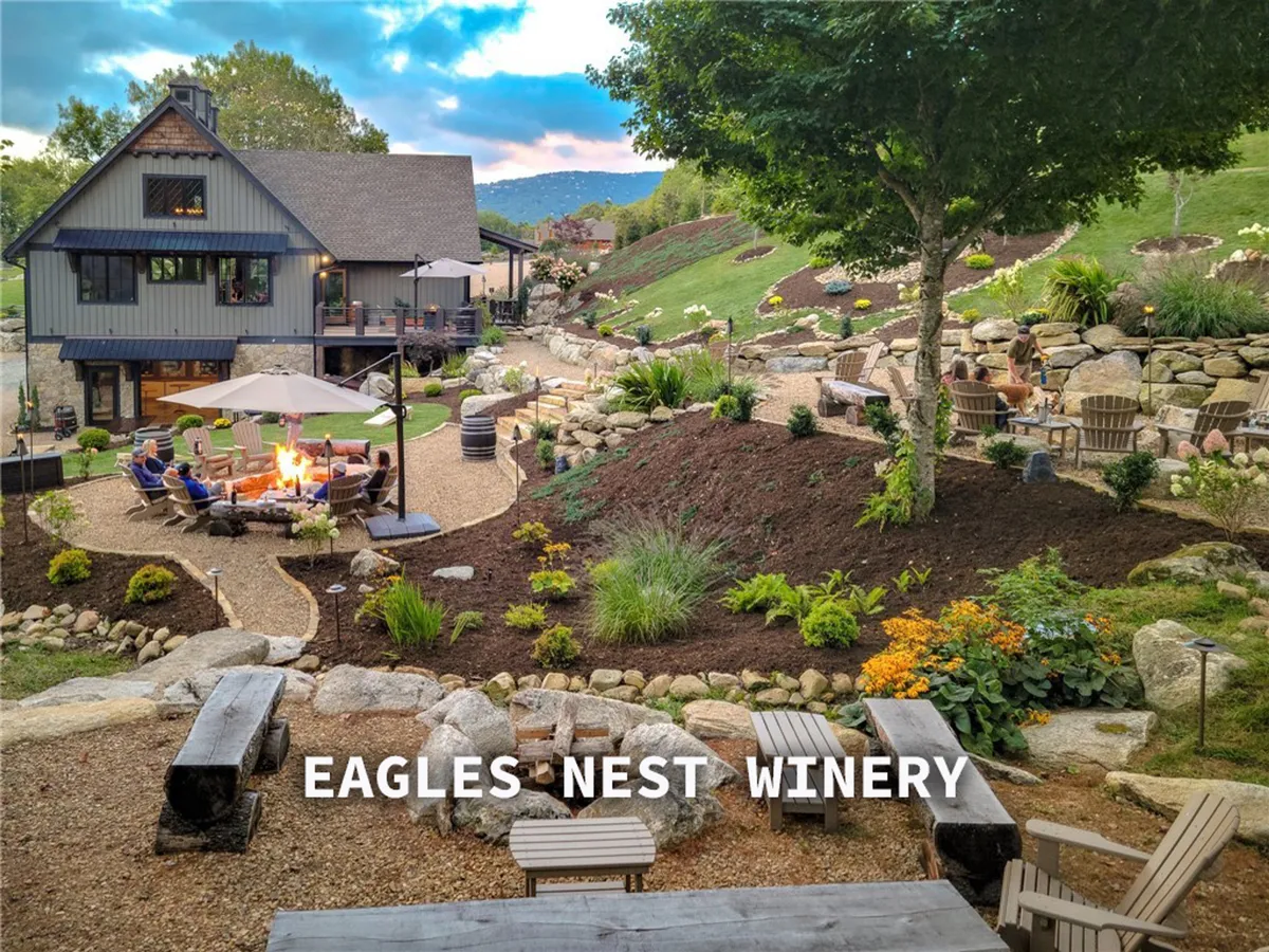 This image features an outdoor fire pit area surrounded by comfortable seating, lush landscaping, and a pathway leading to the winery building in the background. The relaxed setup suggests socializing, wine tasting, and enjoying evenings outdoors with friends or family.