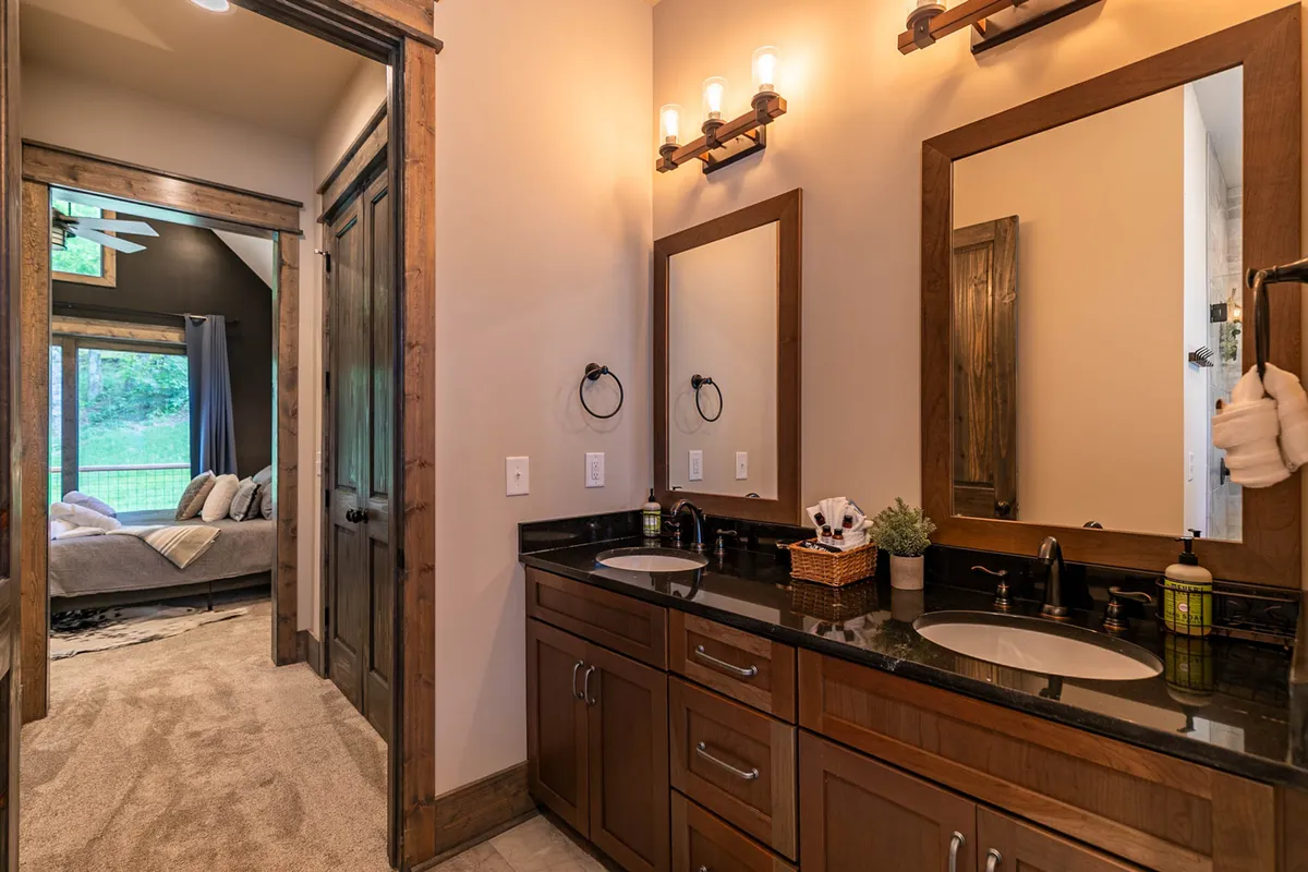 A stylish ensuite bathroom with double vanity sinks, modern light fixtures, mirrors, wooden cabinetry, and a doorway leading to the bedroom. The calm, functional design is perfect for morning routines and self-care.