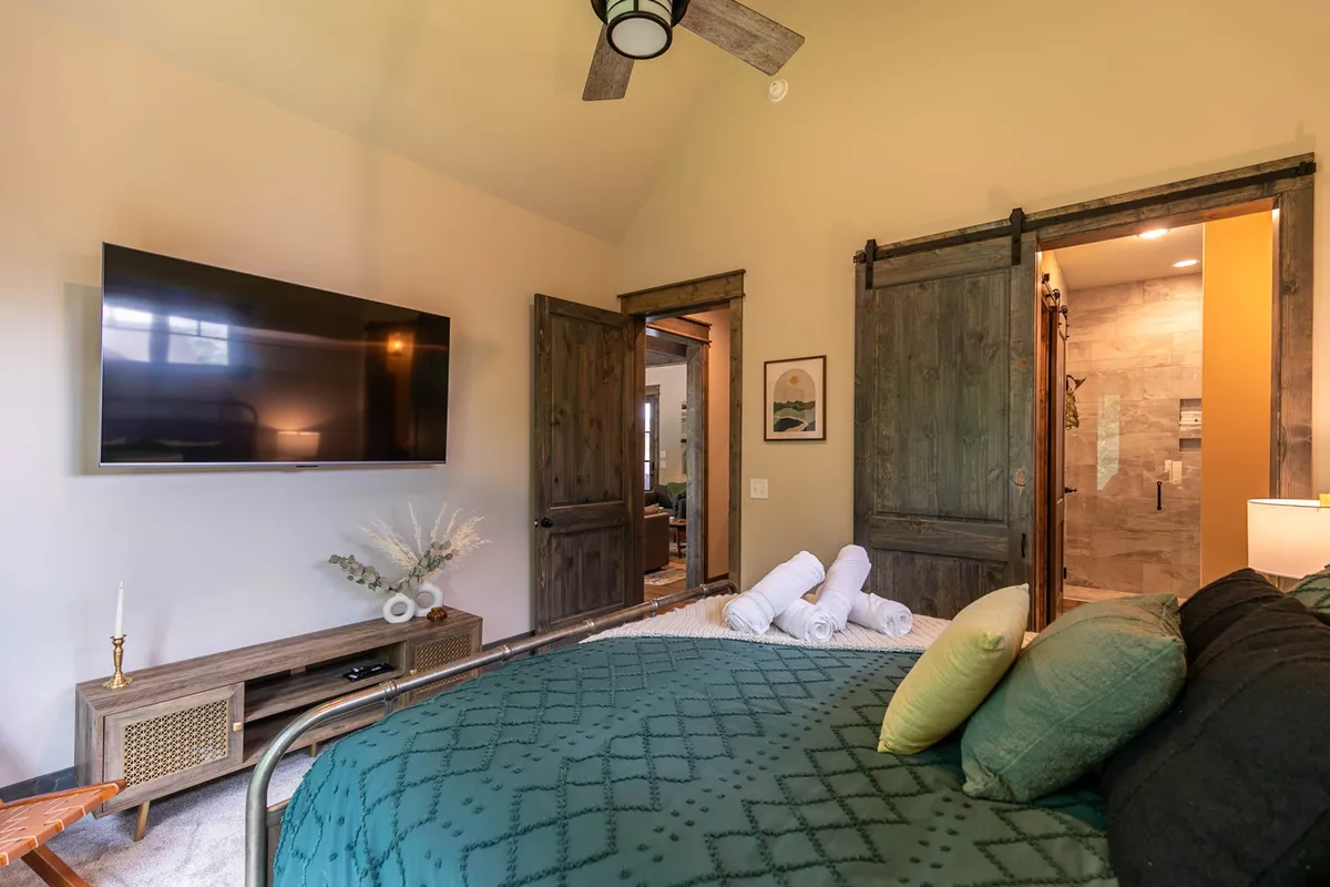 A comfortable king bedroom with a green quilted bed, television, visible bathroom door, and wooden furniture. The space blends comfort and functionality, allowing guests to enjoy entertainment or transition easily to ensuite relaxation.