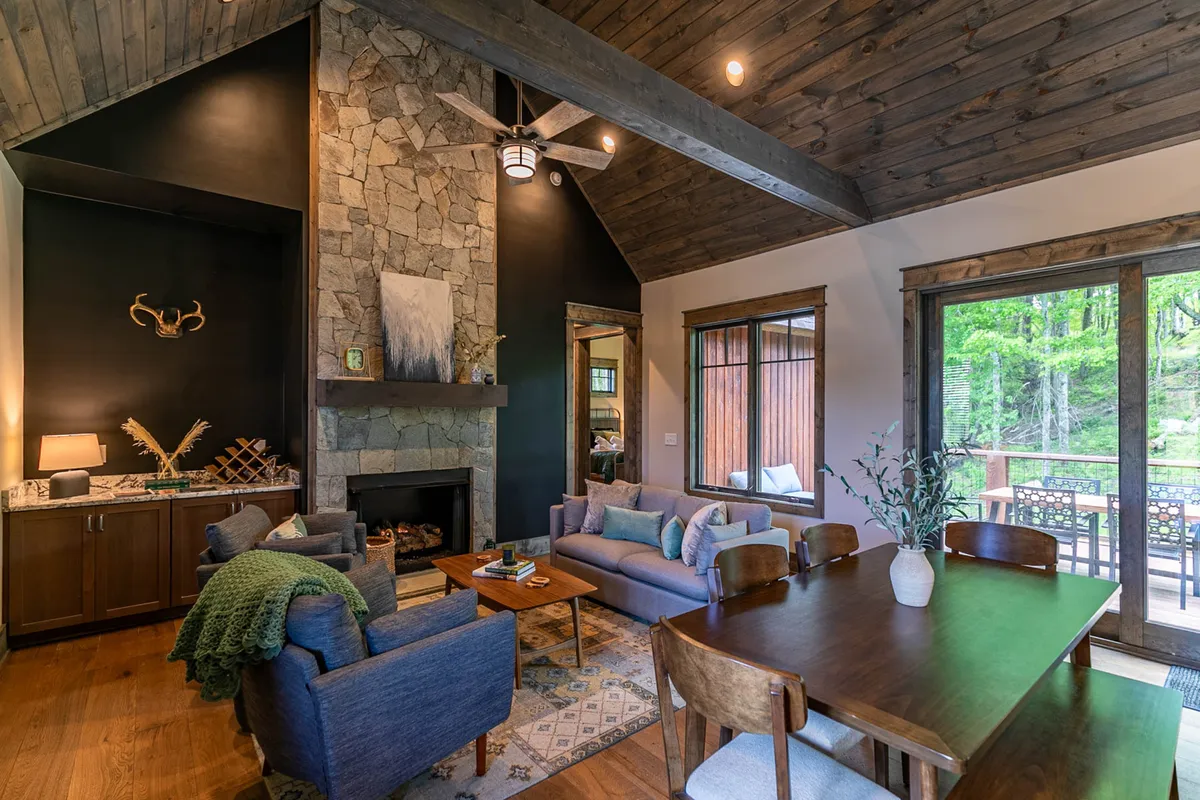 A welcoming interior scene featuring a striking stone fireplace as the focal point, flanked by large windows that let in natural light. There is a spacious wooden dining table set for gathering, comfortable seating in the living area, and wood-planked vaulted ceilings that add rustic charm and openness to the space.