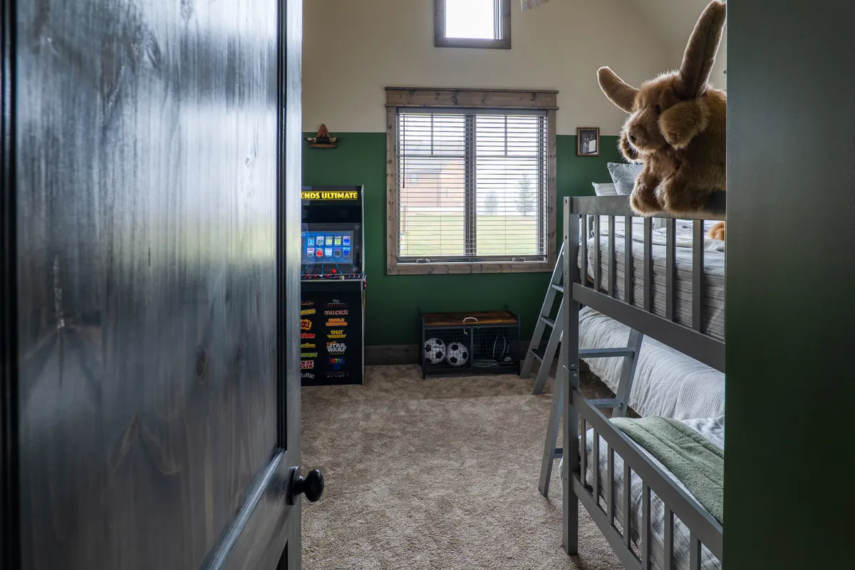 A cheerful kids' bunk room with colorful twin-over-twin bunk beds, a window letting in natural light, a stuffed animal on the bedding, and a retro arcade machine in the corner. The decor is playful and inviting, perfect for sleepovers and games.