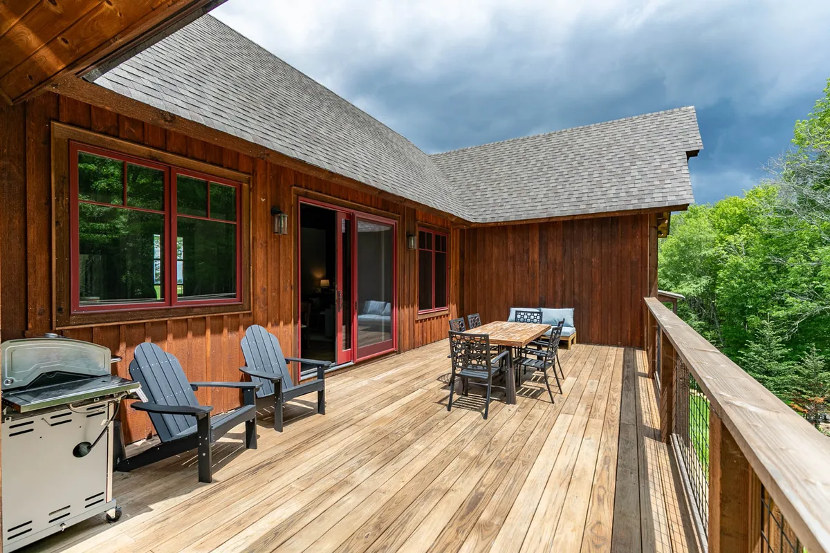 A wooden deck set up with a dining table, grill, and Adirondack chairs, surrounded by nature. The space is ideal for outdoor dining, barbecue nights, or relaxed socializing while enjoying sunset views.