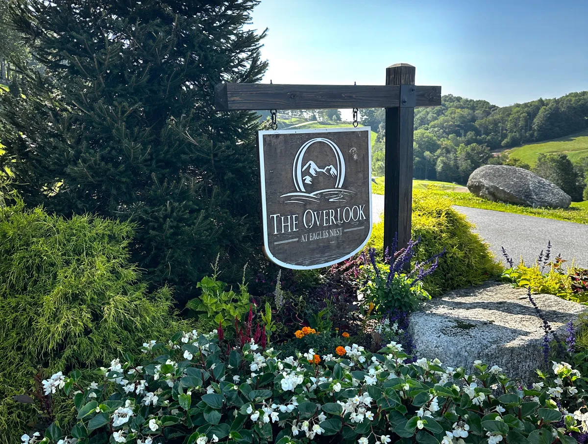 This exterior shot features welcoming signage for 'The Overlook', surrounded by vibrant floral arrangements and set against a natural landscape backdrop. The scene implies immediate access to nature, trailheads, and woodland areas, inviting guests to step right into adventure.