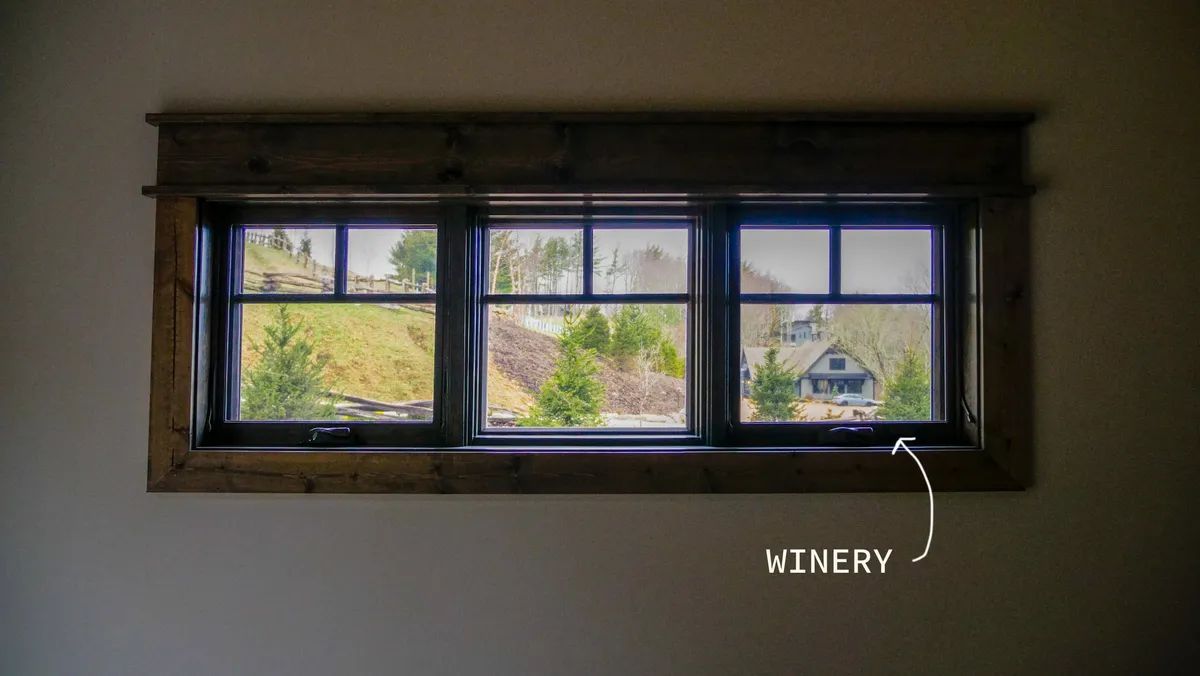 From the interior, this image showcases a window framing a tranquil view of the winery and surrounding greenery. The inviting setting suggests enjoying a glass of wine while taking in the landscape, blending indoor comfort with the beauty just outside.