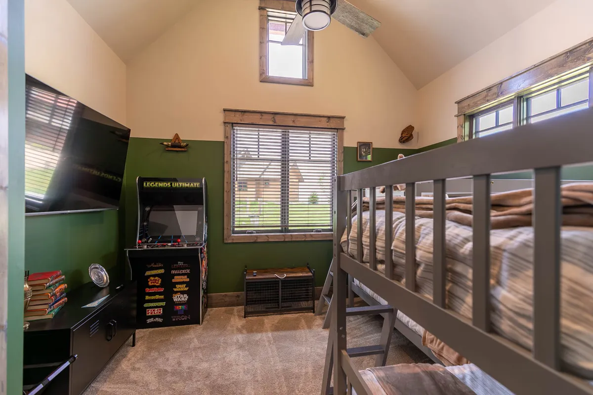A vibrant bunk room featuring two twin-over-twin bunk beds, a retro arcade machine, flat-screen smart TV, bookshelf filled with games and books, and cozy seating areas. The space is arranged for kids to relax, play video games, read, or enjoy time with friends, all in a lively and fun setting.